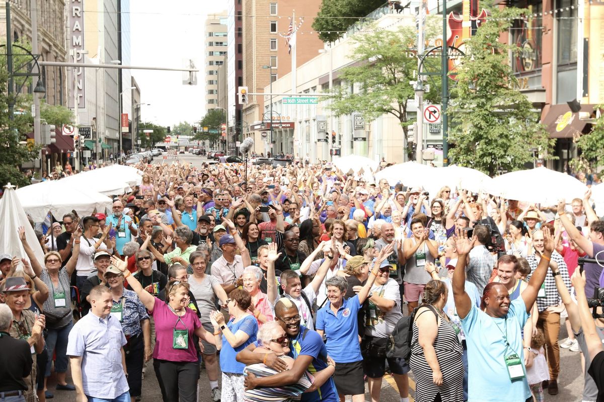 A large, diverse crowd of people gathers in a city street, raising their arms and cheering in a festive, celebratory atmosphere; tall buildings and green trees line the background.