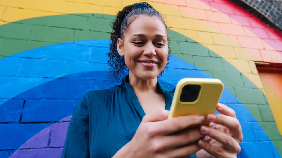 A woman standing in front of a mural of a rainbow, looking down at her phone and smiling.