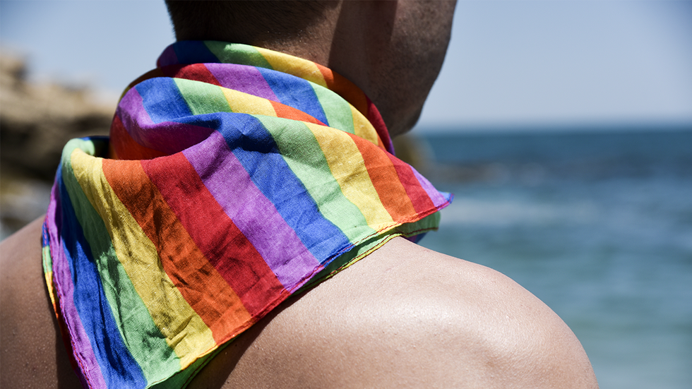 a young caucasian man seen from behind with a rainbow patterned handkerchief tied around his neck in front of the ocean