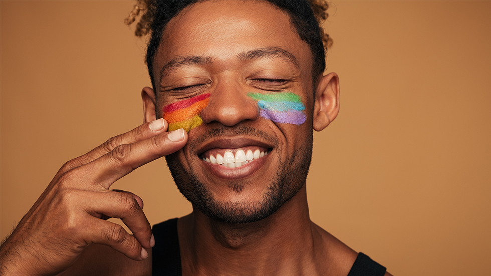 Cheerful African American gay smiling with closed eyes and painting rainbow on cheeks during LGBT pride event against brown background