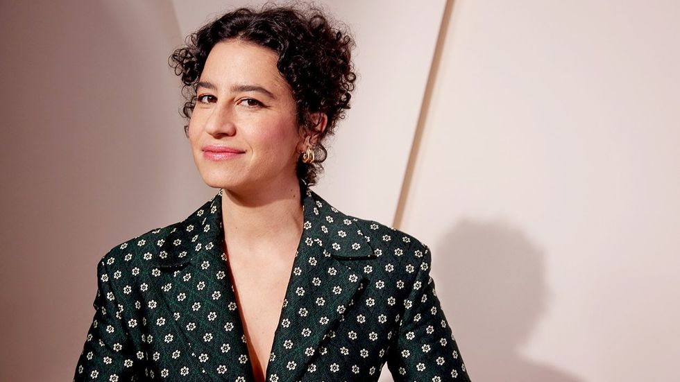 Comedian Ilana Glazer, a 37-year-old Ashkenazi Jewish person with light skin, curly brown hair, and brown eyes, against a very light pink backdrop in a green blazer with a floral pattern. 