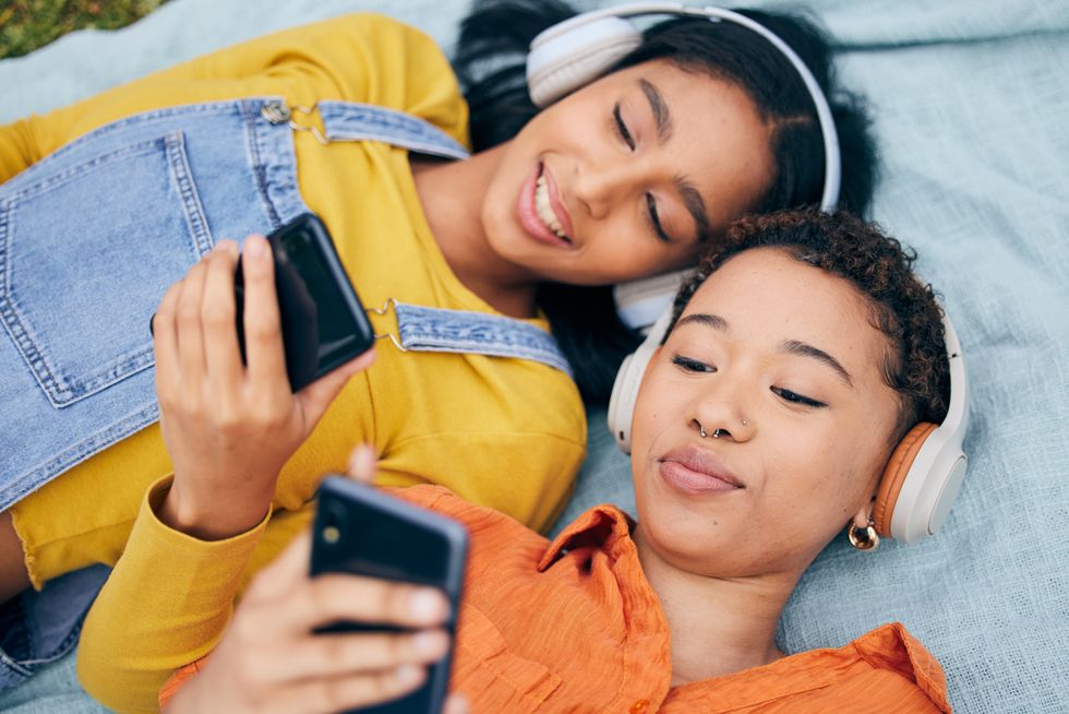 Headphones, phone and lgbt couple on picnic on grass, relax, technology and streaming service in nature. Cellphone, listening to music and happy lesbian women on blanket in garden together from above