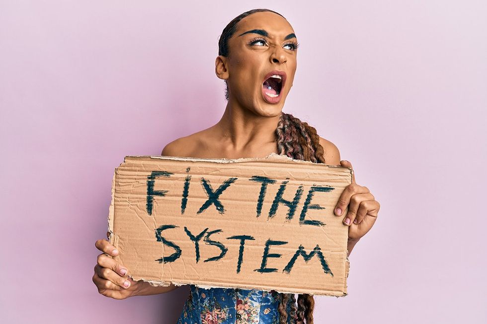 Hispanic man wearing make up and long hair holding fix the system banner cardboard angry and mad screaming frustrated and furious, shouting with anger. rage and aggressive concept.