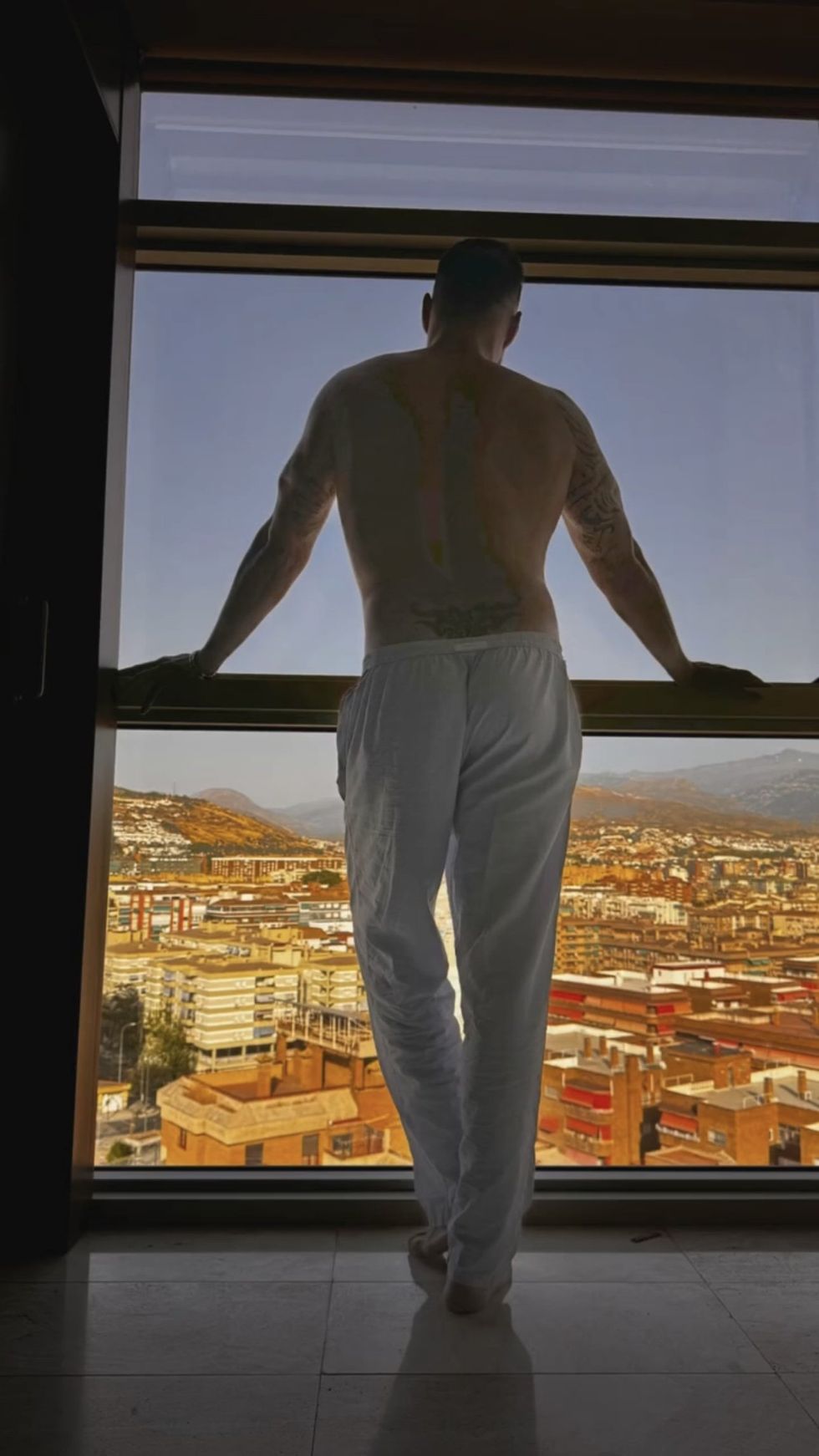 Ricky Martin from behind, shirtless and in loose white pants, overlooking a city from a window.