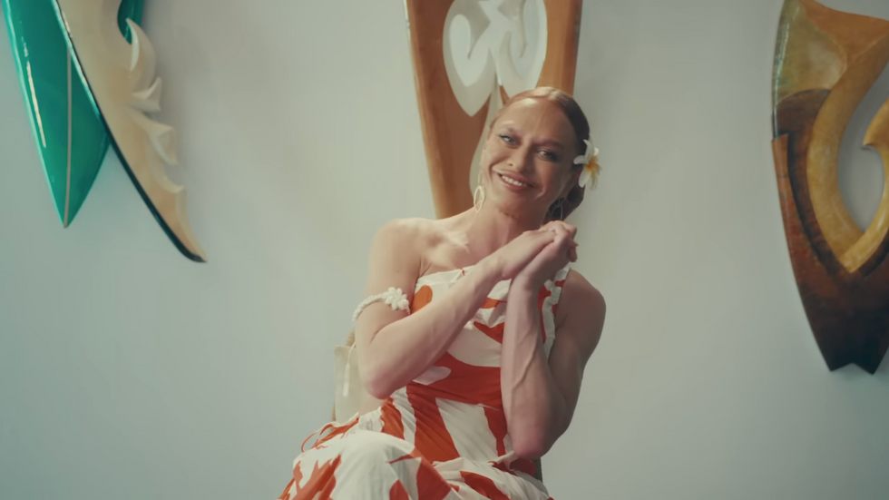 Sasha Colby in an orange and white dress with a white flower in her slicked back hair, getting interviewed during the HOME STRETCH documentary.