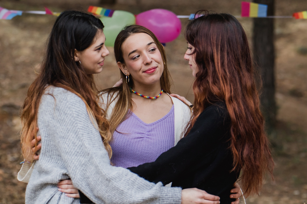 Three women stand as some sort of pride event. They're in a triangle, all loosely wrapped in one another's arms.