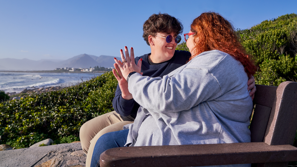 Two lesbians sitting on a bench by the coast. They're looking lovingly at each other and have their palms pressed against one another.