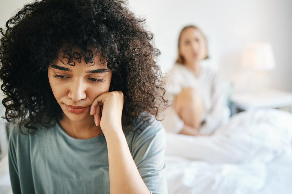 Two women in a bedroom. One, in the back and out of focus, sits at the head of the bed staring at the woman in focus. The woman in focus sits with her head in her hand, looking forlorn.