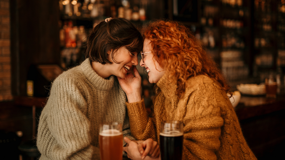 Two women in knitwear who are laughing with one another and sharing a beer