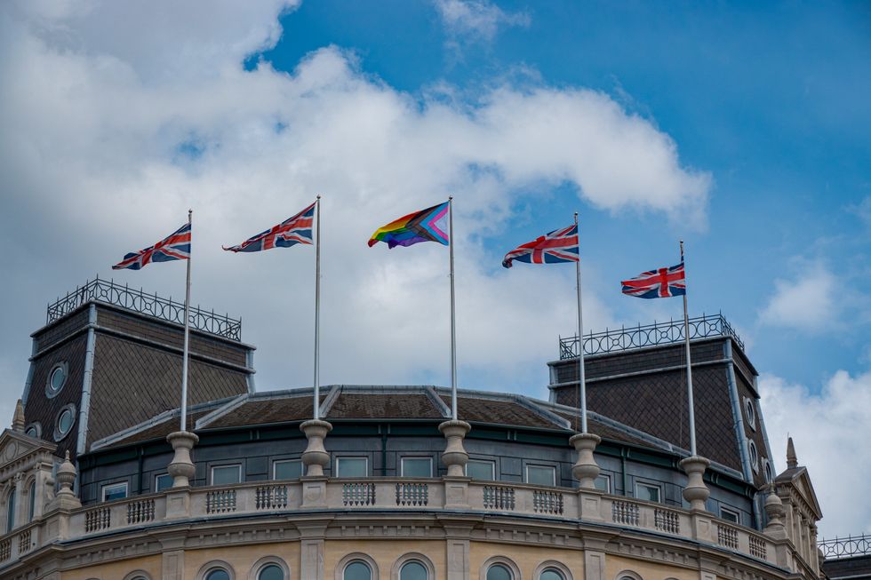 Union Jack and Pride flags flying side by side atop an old government building in London, drawing attention to an existing societal issue of gender orientation and symbolizing diversity and inclusion.