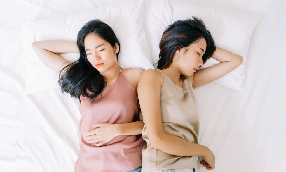 Upset young Asian lesbian couple having a disagreement lying side by side in bed facing in opposite directions ignoring one another in the bedroom. Negative emotions concept