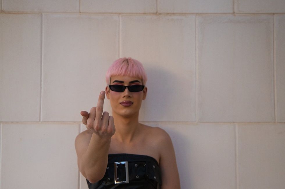 Young gay guy with pink hair and makeup and sunglasses is leaning on a white wall making the finger gesture of fuck you. Concept of equality and lgbtq rights